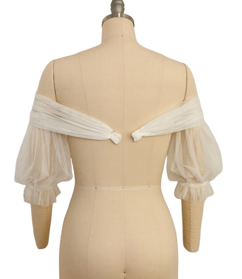 May include: White off-the-shoulder tulle bridal bolero with puffed sleeves and a gathered detail at the wrist.