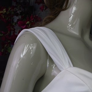 May include: A white dress featuring a wide, gathered shoulder strap and a fitted bodice. The garment is displayed on a glossy, light-coloured mannequin, highlighting the smooth texture and clean lines of the white fabric against the form.