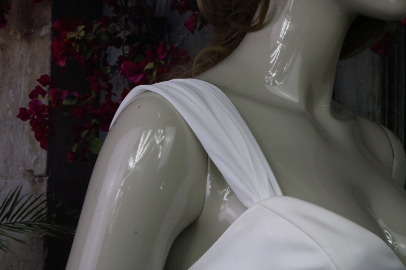 May include: A white dress featuring a wide, gathered shoulder strap and a fitted bodice. The garment is displayed on a glossy, light-coloured mannequin, highlighting the smooth texture and clean lines of the white fabric against the form.