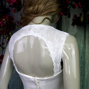 Könnte beinhalten: Ein weißes Brautkleid mit einem Spitzenoberteil und einem enganliegenden Rock. Das Oberteil hat einen tiefen Rücken mit einer Spitzenüberlagerung. Das Kleid wird hinten mit Knöpfen geschlossen.