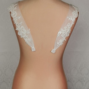 May include: A pair of white lace bridal shoulder straps with delicate floral patterns and sequins. The straps are attached to a nude-coloured mannequin.