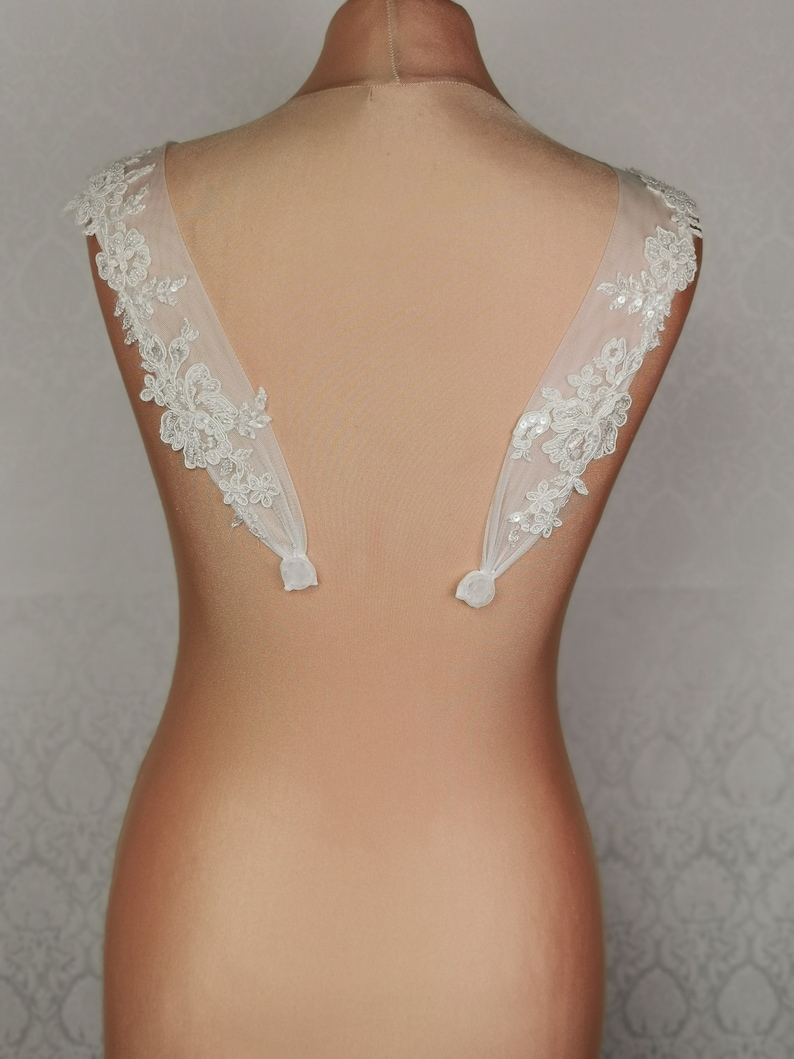 May include: A pair of white lace bridal shoulder straps with delicate floral patterns and sequins. The straps are attached to a nude-coloured mannequin.