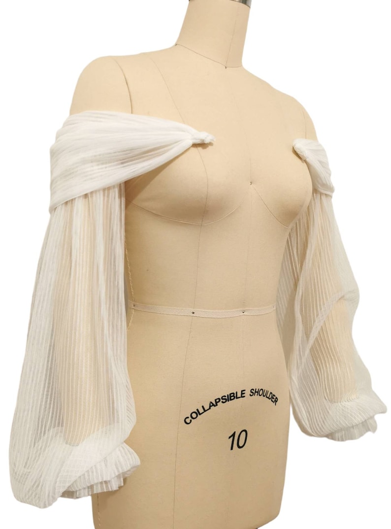 Puede incluir: Mangas blancas transparentes de hombros descubiertos con un detalle fruncido en la parte superior e inferior. Las mangas est&aacute;n hechas de una tela ligera y tienen un aspecto delicado y fluido. "COLLAPSIBLE SHOULDER 10" est&aacute; escrito en el maniqu&iacute;.