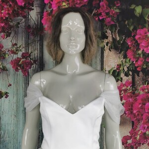 May include: White off-the-shoulder top with a sweetheart neckline and sheer tulle sleeves. The top is made of a smooth, structured fabric.