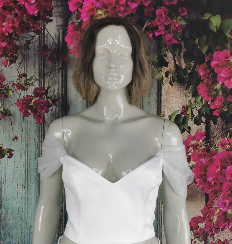 May include: White off-the-shoulder top with a sweetheart neckline and sheer tulle sleeves. The top is made of a smooth, structured fabric.