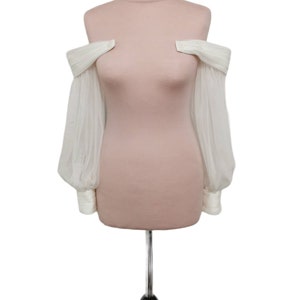 Puede incluir: Una blusa blanca de hombros descubiertos con mangas largas y abullonadas transparentes. La blusa est&aacute; en un maniqu&iacute;.