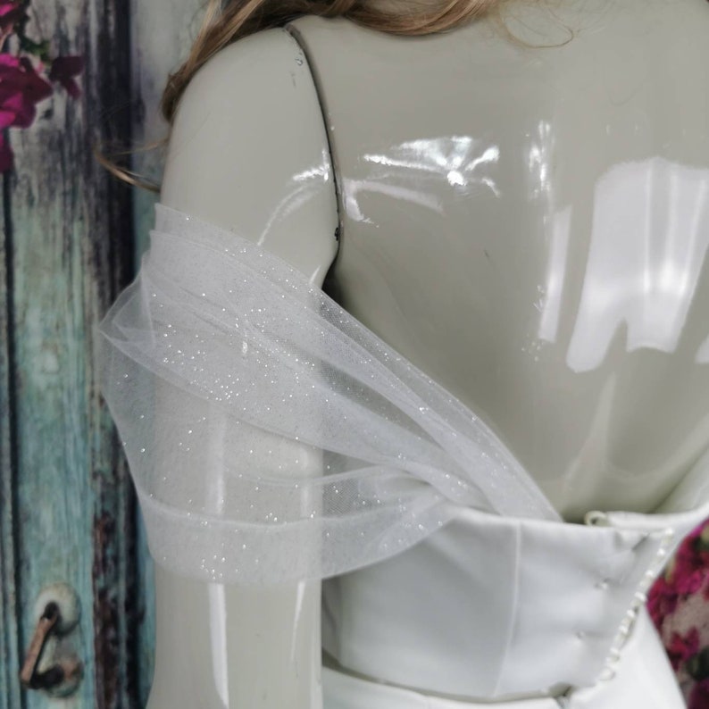 May include: White off-the-shoulder wedding dress with a tulle overlay featuring sparkly embellishments.