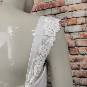 May include: White wedding dress with a lace detail on the straps. The straps are made of a sheer fabric and are adorned with delicate white lace.