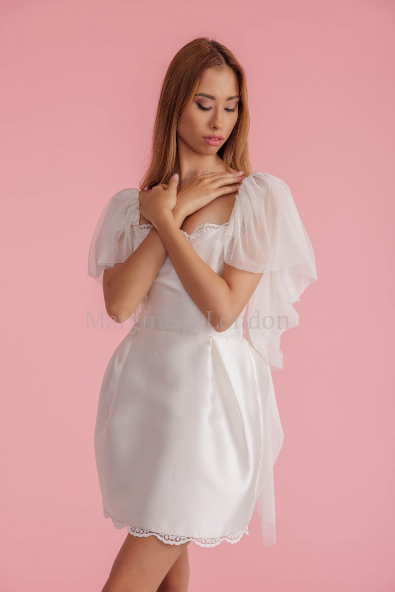 Puede incluir: Un vestido blanco de sat&eacute;n con un borde de encaje y mangas abullonadas transparentes. El vestido tiene un escote de coraz&oacute;n y un corpi&ntilde;o ajustado. La modelo lleva el vestido sobre un fondo rosa.
