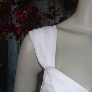 May include: A white dress featuring a wide, gathered shoulder strap that connects to a smooth, solid white bodice. The fabric appears opaque and matte, draping over the shoulder of a mannequin to showcase the construction of the strap.
