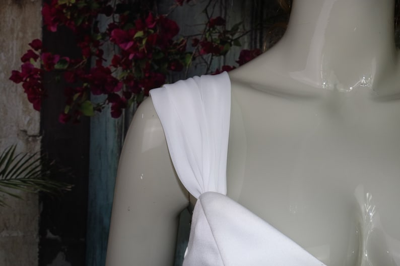 May include: A white dress featuring a wide, gathered shoulder strap that connects to a smooth, solid white bodice. The fabric appears opaque and matte, draping over the shoulder of a mannequin to showcase the construction of the strap.