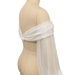 Puede incluir: Top de novia blanco de hombros descubiertos con mangas largas, transparentes y plisadas. El top se ata en la espalda con un lazo.