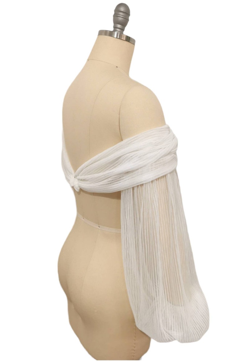 Puede incluir: Top de novia blanco de hombros descubiertos con mangas largas, transparentes y plisadas. El top se ata en la espalda con un lazo.