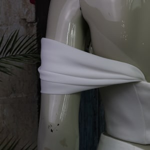 May include: A white off-the-shoulder garment featuring a smooth, matte fabric bodice with a draped, horizontal sleeve detail that wraps around the upper arm, displayed on a glossy, light-coloured mannequin torso.