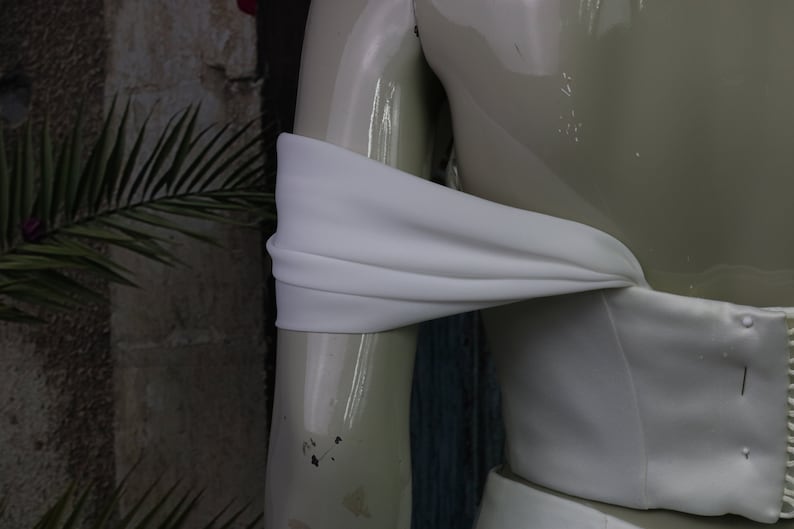 May include: A white off-the-shoulder garment featuring a smooth, matte fabric bodice with a draped, horizontal sleeve detail that wraps around the upper arm, displayed on a glossy, light-coloured mannequin torso.
