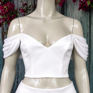 May include: A white crop top featuring a V-neckline and off-the-shoulder sleeves with gathered fabric detailing. The garment is displayed on a mannequin, showcasing a smooth, solid white fabric construction with a fitted bodice design.