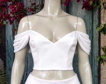 May include: A white crop top featuring a V-neckline and off-the-shoulder sleeves with gathered fabric detailing. The garment is displayed on a mannequin, showcasing a smooth, solid white fabric construction with a fitted bodice design.