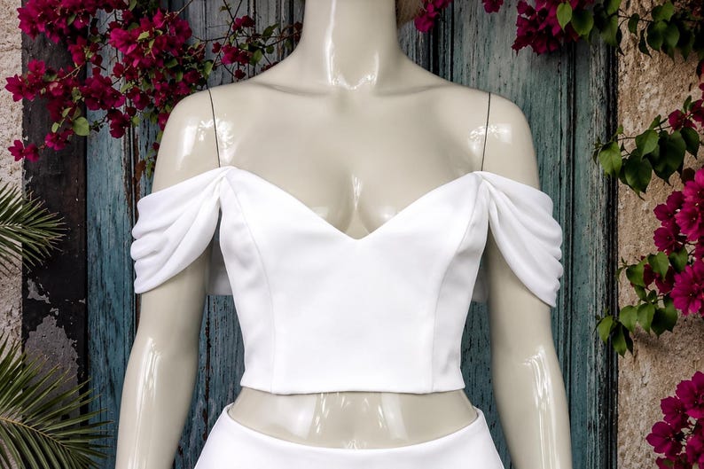 May include: A white crop top featuring a V-neckline and off-the-shoulder sleeves with gathered fabric detailing. The garment is displayed on a mannequin, showcasing a smooth, solid white fabric construction with a fitted bodice design.