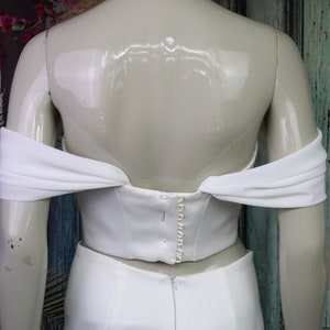May include: A white off-the-shoulder corset top featuring draped fabric sleeves and a vertical row of small white buttons along the centre back closure, displayed on a mannequin against a weathered teal wooden door.