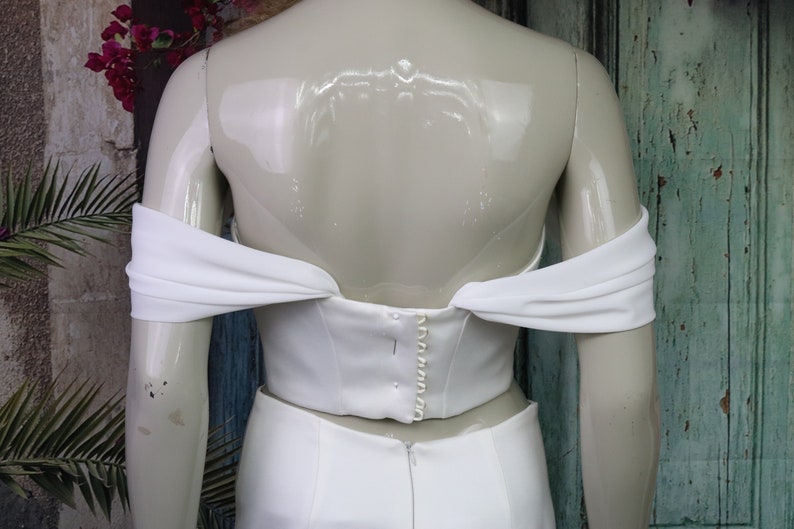 May include: A white off-the-shoulder corset top featuring draped fabric sleeves and a vertical row of small white buttons along the centre back closure, displayed on a mannequin against a weathered teal wooden door.