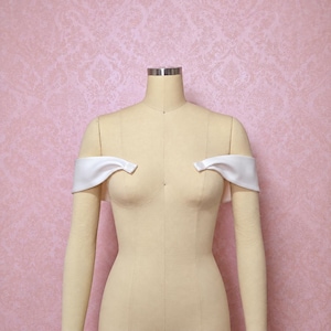 May include: A pair of white satin off-the-shoulder sleeves displayed on a beige dress form. The sleeves feature a smooth, lustrous finish and are positioned to drape across the upper arms, creating a clean, structured look for formal attire.