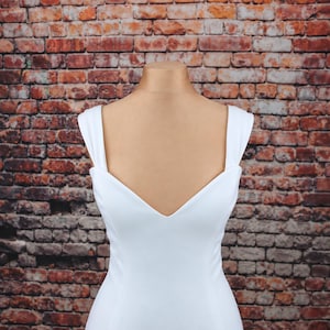 May include: A solid white sleeveless dress featuring a deep V-neckline and wide shoulder straps. The garment is displayed on a mannequin, showcasing a clean, smooth fabric texture and a fitted bodice design that extends downward.
