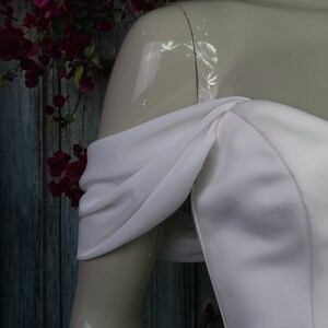 May include: A white off-the-shoulder dress featuring draped fabric sleeves that wrap around the upper arms. The bodice is constructed from a smooth, solid white material with a structured neckline that sits across the chest and shoulders.