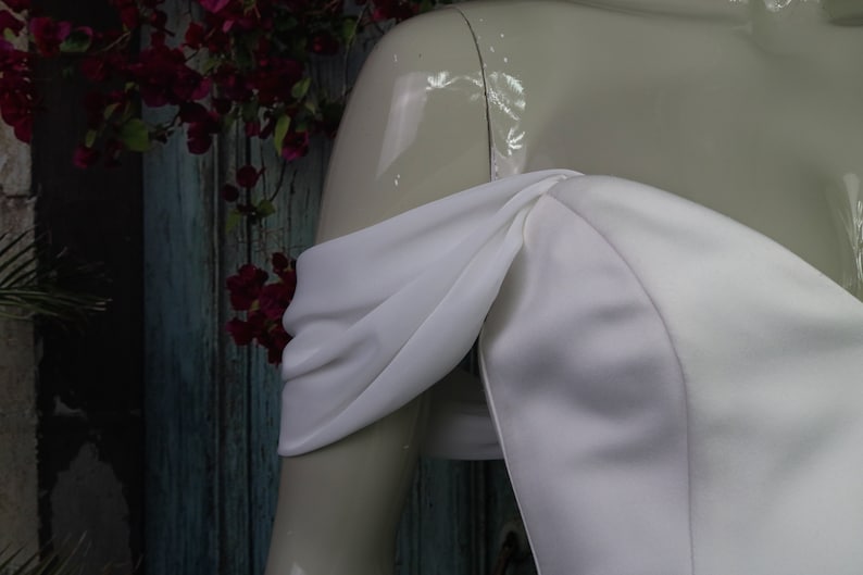 May include: A white off-the-shoulder dress featuring draped fabric sleeves that wrap around the upper arms. The bodice is constructed from a smooth, solid white material with a structured neckline that sits across the chest and shoulders.