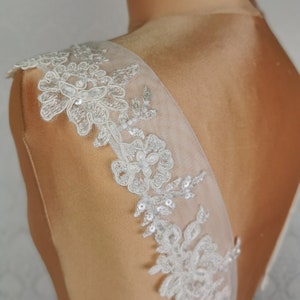 May include: White lace bridal sash with delicate floral patterns and sequins. The sash is shown on a dress form and is designed to be worn on the back of a wedding dress.