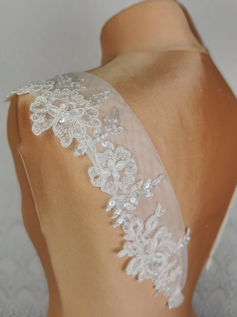 May include: White lace bridal sash with delicate floral patterns and sequins. The sash is shown on a dress form and is designed to be worn on the back of a wedding dress.