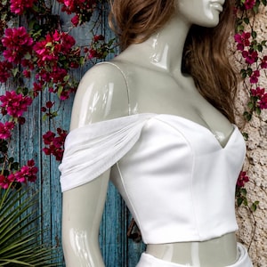 May include: A white off-the-shoulder crop top featuring a sweetheart neckline and draped, sheer fabric sleeves. The garment is fitted through the bodice and is displayed on a mannequin, showcasing a clean, structured design.