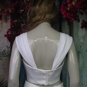 May include: A white sleeveless top featuring wide, draped shoulder straps and a deep V-shaped back. The garment includes a central vertical row of small, round white buttons along the back closure, providing a structured fit.