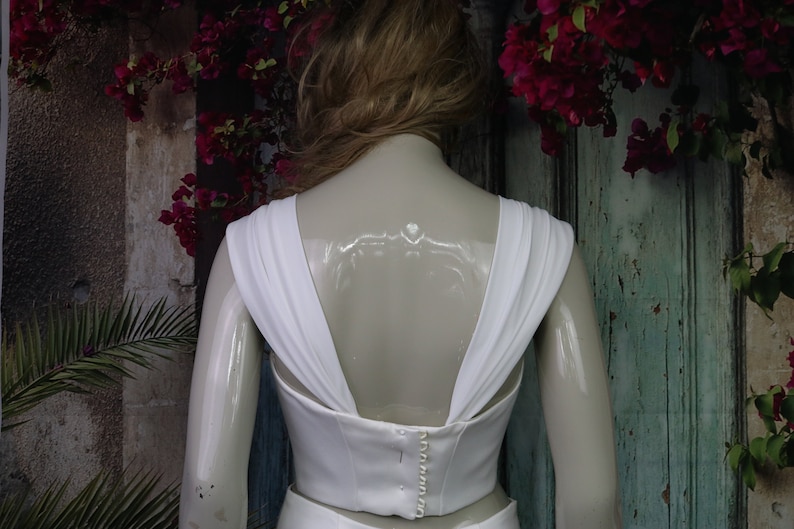 May include: A white sleeveless top featuring wide, draped shoulder straps and a deep V-shaped back. The garment includes a central vertical row of small, round white buttons along the back closure, providing a structured fit.