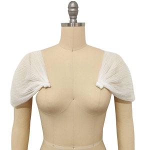 May include: White sheer fabric bridal bolero with short sleeves. The bolero is pleated and has a delicate, feminine look.