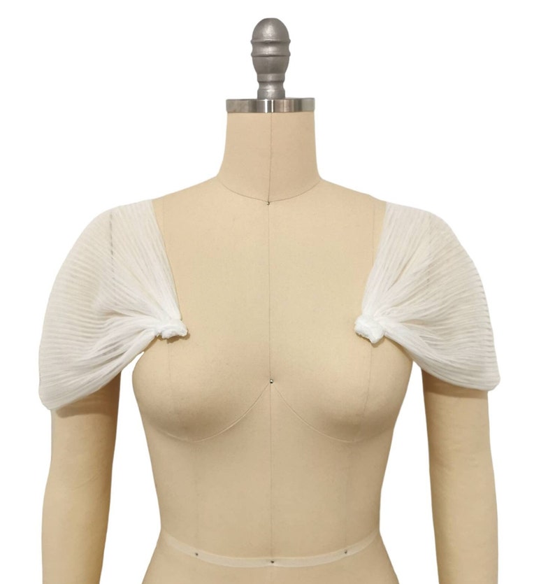 May include: White sheer fabric bridal bolero with short sleeves. The bolero is pleated and has a delicate, feminine look.