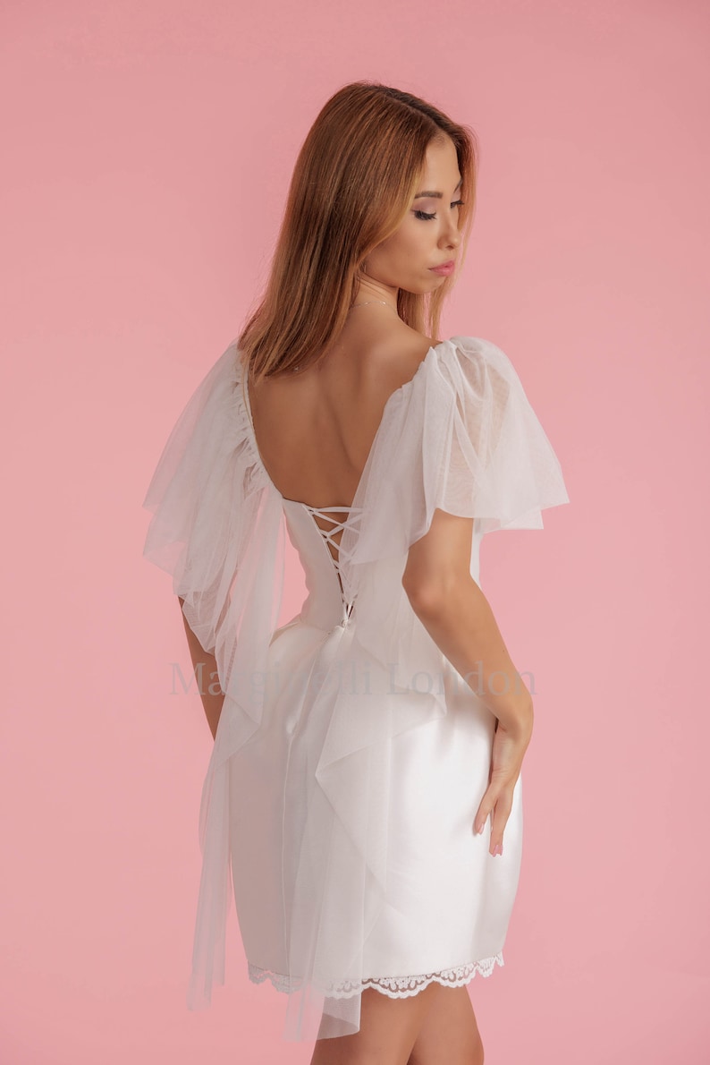 Puede incluir: Un vestido blanco con ribete de encaje y espalda con cordones. El vestido tiene una superposici&oacute;n de tul con mangas con volantes.