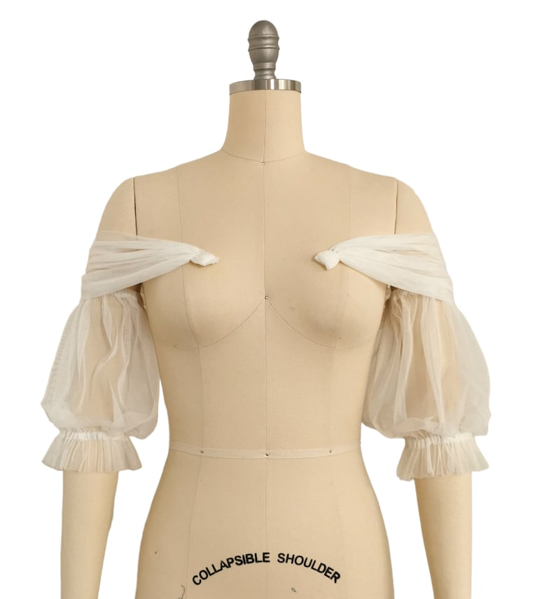 May include: White off-the-shoulder tulle bridal bolero with puffed sleeves and a gathered ruffle trim.