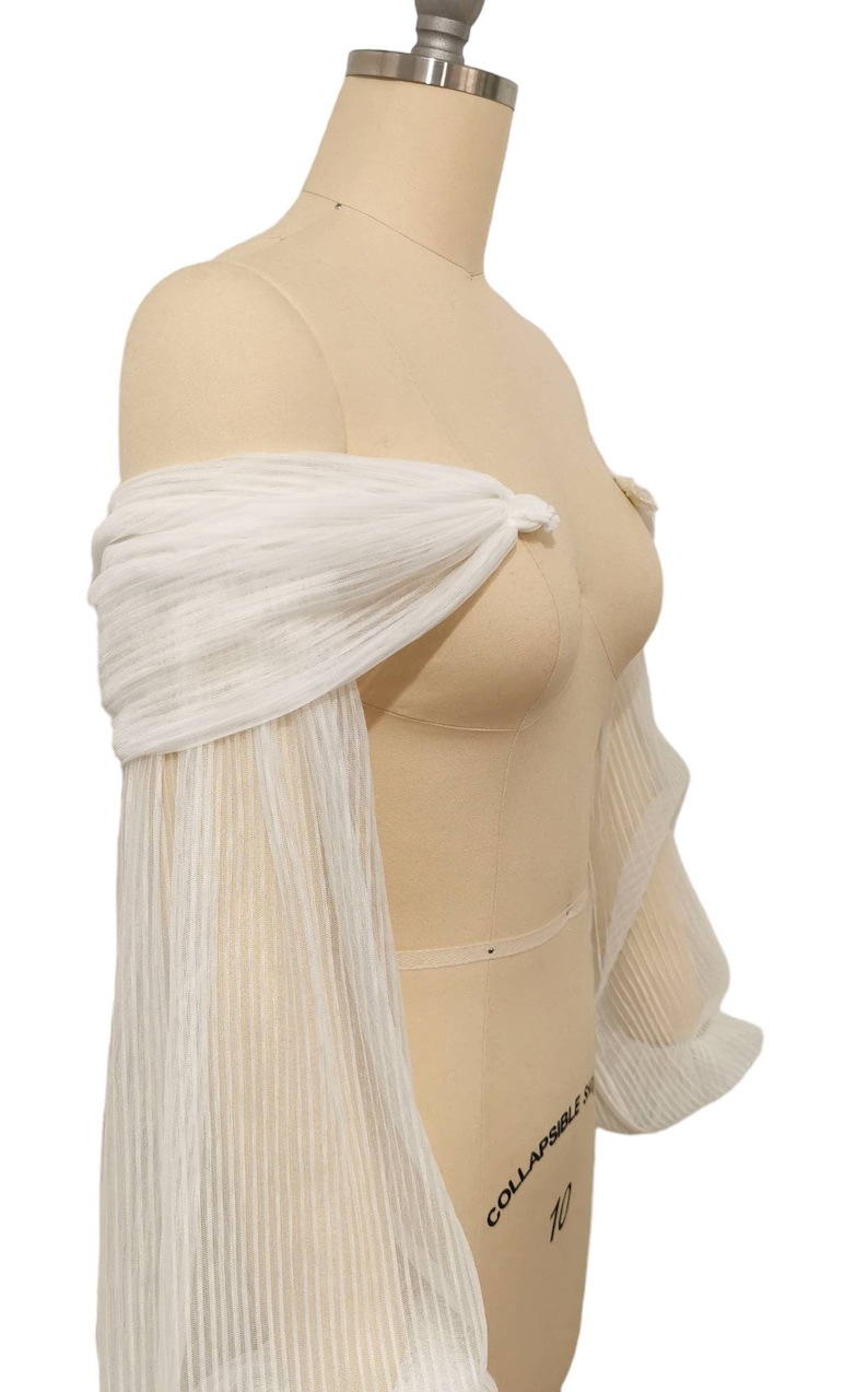 Puede incluir: Bolero de novia blanco, sin hombros, con mangas largas. Las mangas est&aacute;n hechas de una tela transparente y plisada.