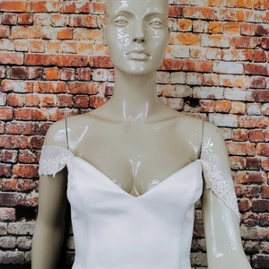 May include: A white off-the-shoulder wedding dress with lace detailing on the straps. The dress has a sweetheart neckline and a fitted bodice.