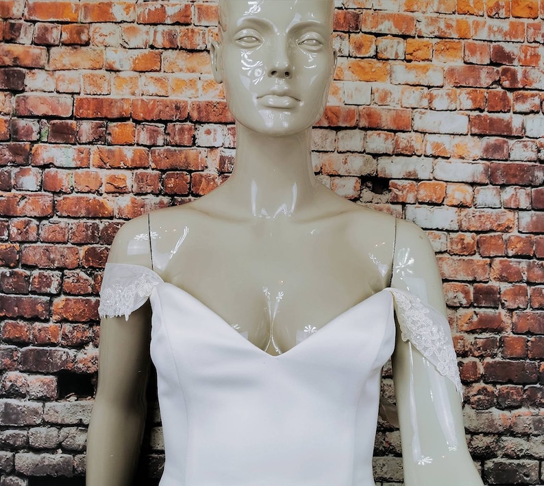 May include: A white off-the-shoulder wedding dress with lace detailing on the straps. The dress has a sweetheart neckline and a fitted bodice.