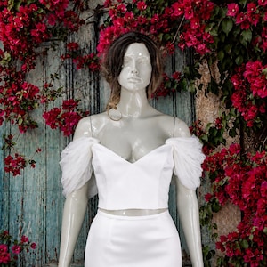 May include: A white two-piece garment featuring a cropped bodice with a sweetheart neckline and sheer, gathered tulle off-the-shoulder sleeves, paired with a matching floor-length skirt on a mannequin.
