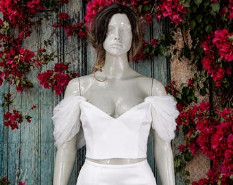 May include: A white two-piece garment featuring a cropped bodice with a sweetheart neckline and sheer, gathered tulle off-the-shoulder sleeves, paired with a matching floor-length skirt on a mannequin.