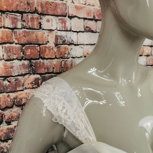 May include: Close-up of a white wedding dress with a lace trim on the shoulder straps.