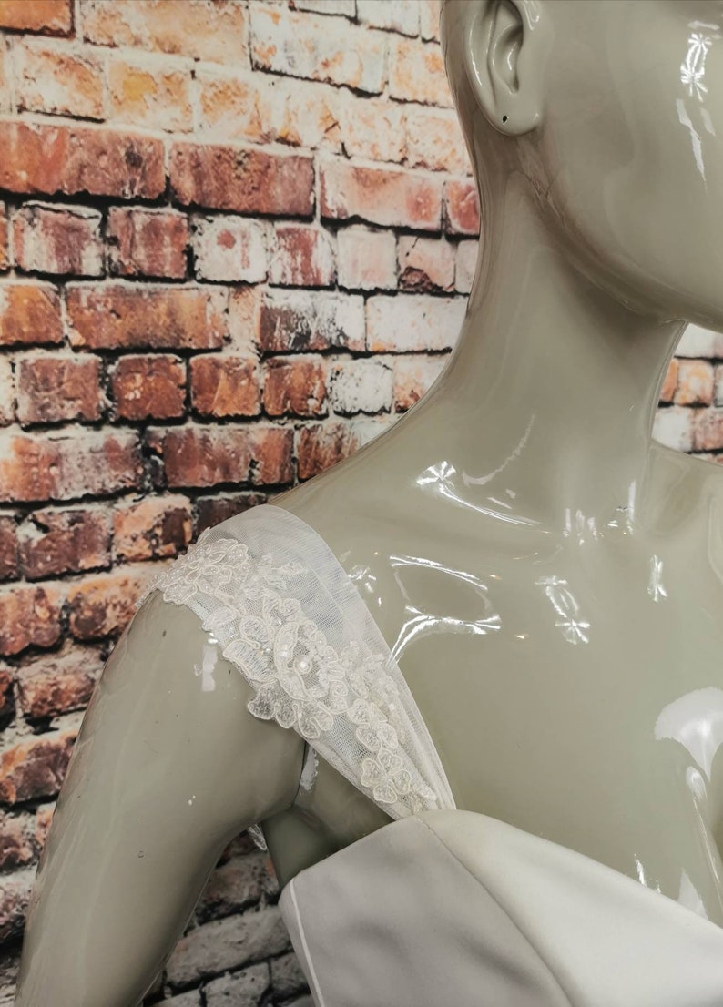 May include: Close-up of a white wedding dress with a lace trim on the shoulder straps.