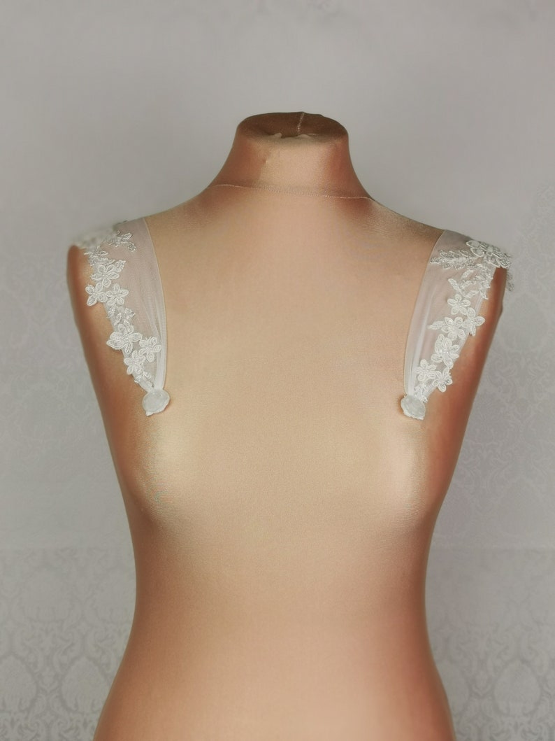 May include: A pair of white tulle and lace bridal shoulder straps with small pearl buttons. The straps are designed to be worn with a strapless dress.