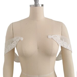 Puede incluir: Bolero de novia blanco sin hombros de encaje con delicado bordado floral. El bolero se muestra en un maniqu&iacute; blanco.