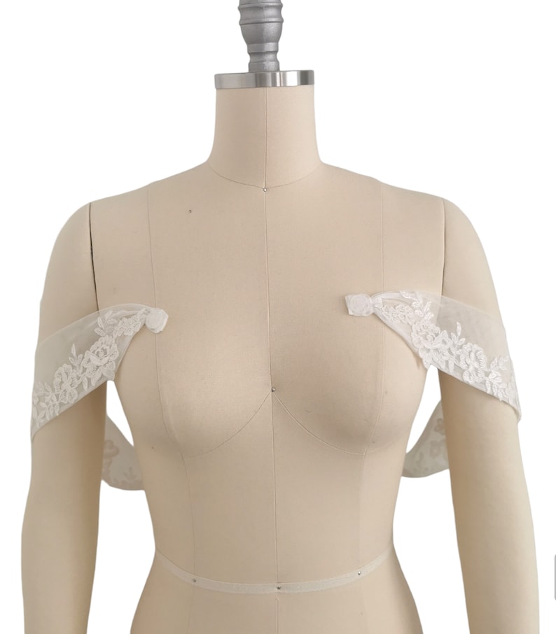 Puede incluir: Bolero de novia blanco sin hombros de encaje con delicado bordado floral. El bolero se muestra en un maniqu&iacute; blanco.