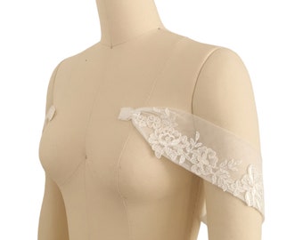 May include: White lace bridal shoulder straps with floral embroidery. The straps are made of a sheer fabric and are designed to be worn over a wedding dress.