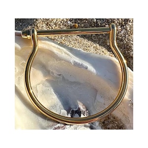May include: A gold-colored, U-shaped bracelet with a straight bar across the top. The bracelet is made of a shiny metal and is resting on a white, textured surface, with sand in the background. The bracelet is a simple, elegant design.