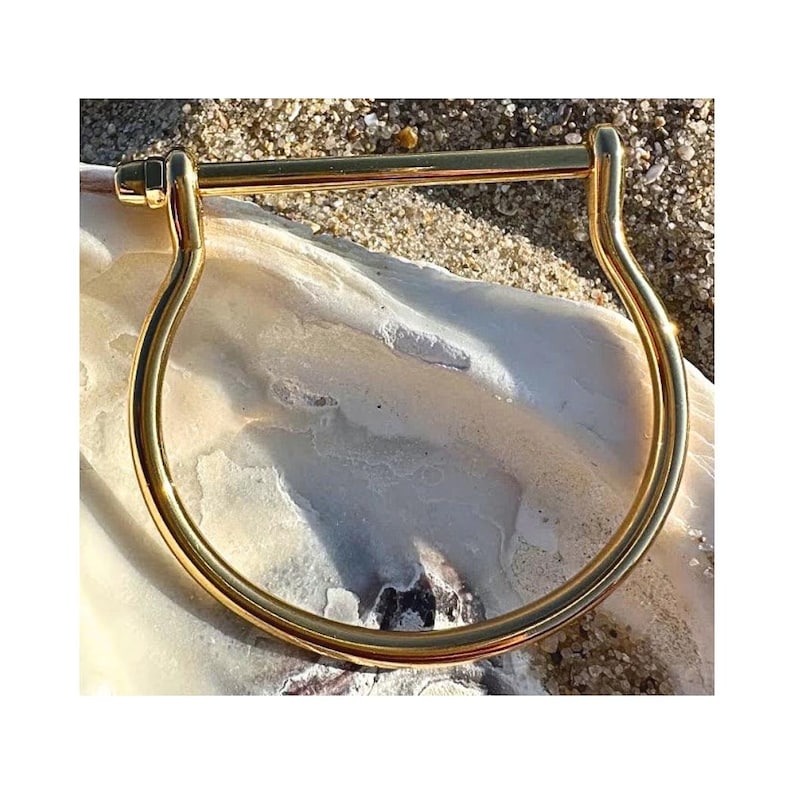 May include: A gold-colored, U-shaped bracelet with a straight bar across the top. The bracelet is made of a shiny metal and is resting on a white, textured surface, with sand in the background. The bracelet is a simple, elegant design.